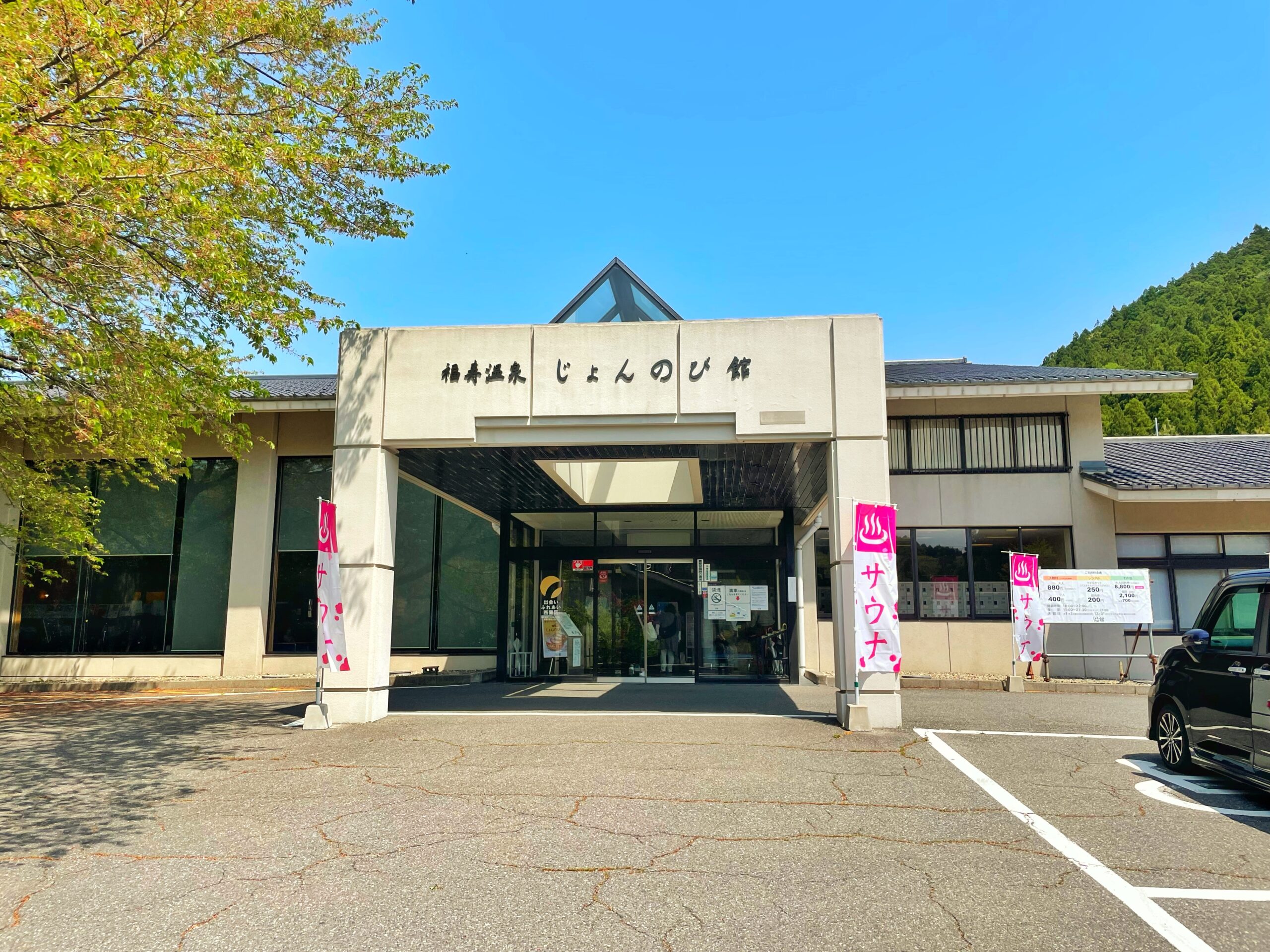 福寿温泉じょんのび館の正面エントランス｜青空と新緑に囲まれたベージュの瓦屋根、入口にピンクのサウナのぼり旗が並ぶ外観｜新潟県新潟市西蒲区