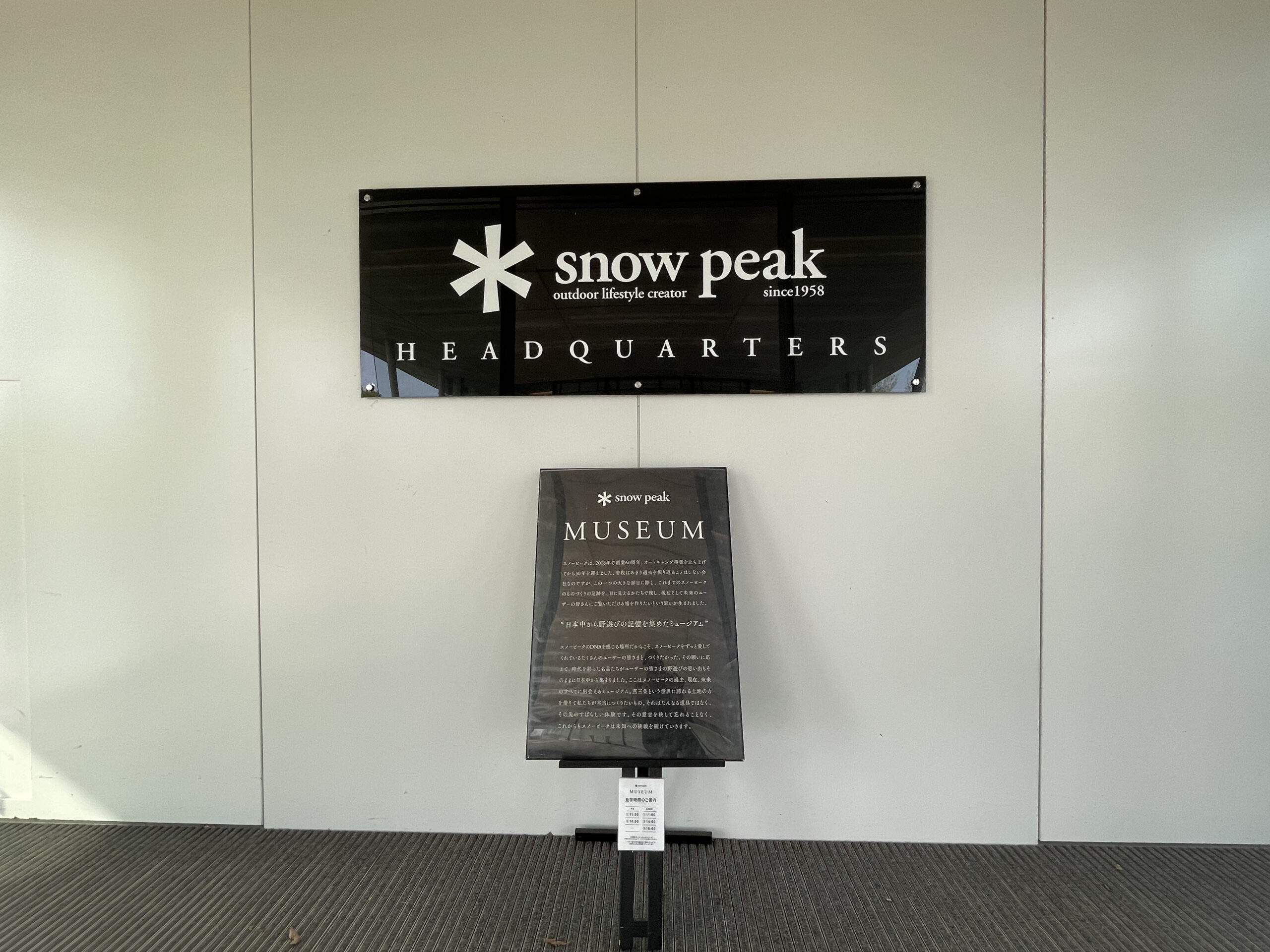 Snow Peak FIELD SUITE SPA HEADQUARTERSのミュージアム案内｜白壁に黒いロゴ看板とMUSEUMのスタンド看板が置かれた館内通路｜新潟県三条市