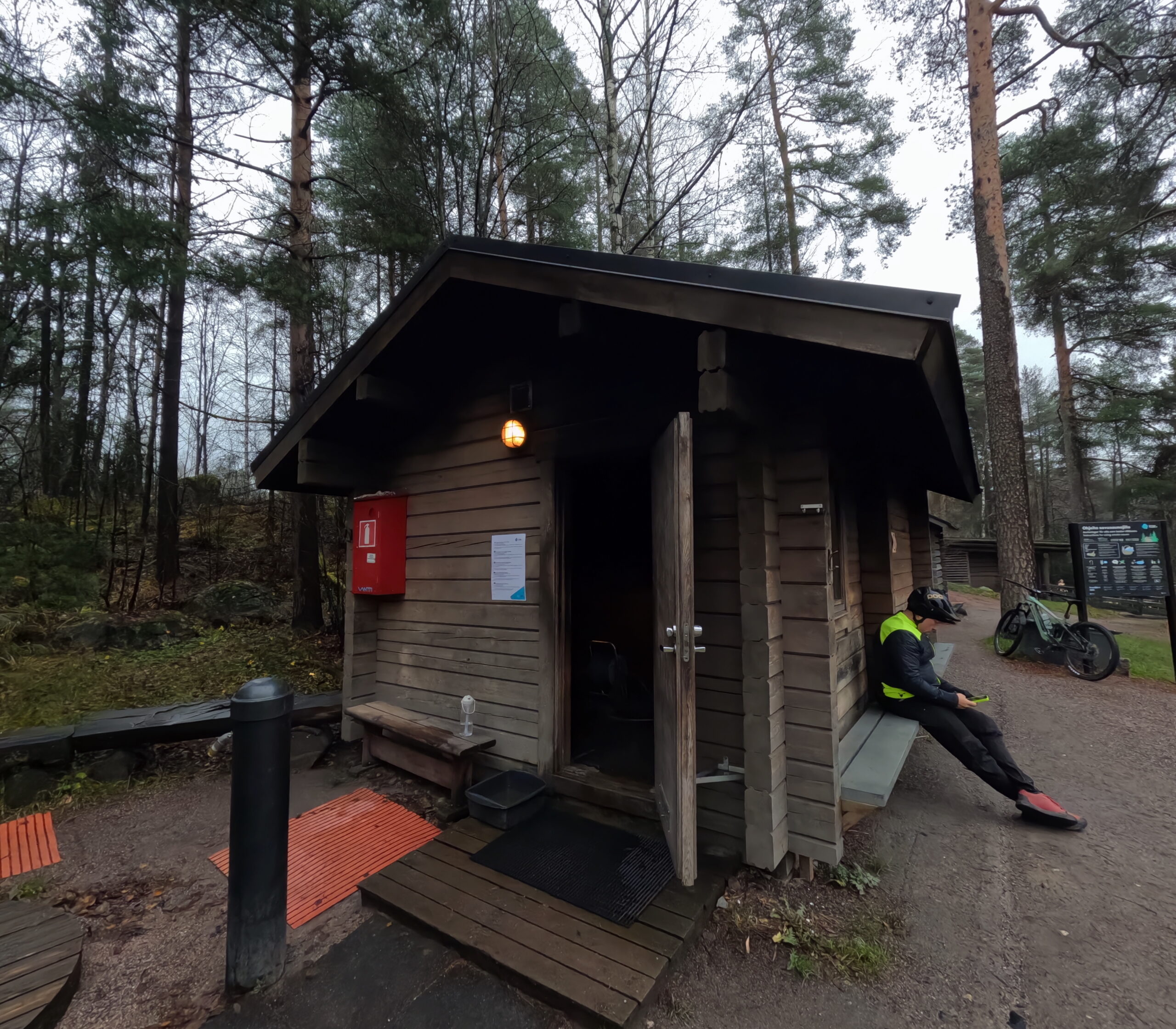 Kuusijärvi伝統的小型スモークサウナ｜丸太造りの素朴な外観、赤い消火箱と外灯、訪問者が休憩する秋の森｜ヴァンター市・フィンランド