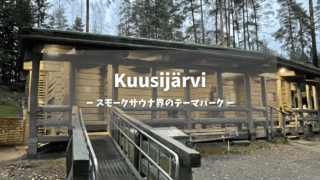 Kuusijärvi（クーシャルヴィ）大型スモークサウナ棟｜温かい照明が灯る丸太造りの木製建築、手すり付きスロープと広いベランダ、針葉樹の森に囲まれた夕暮れの雰囲気｜スモークサウナ界のテーマパーク｜ヴァンター市・フィンランド