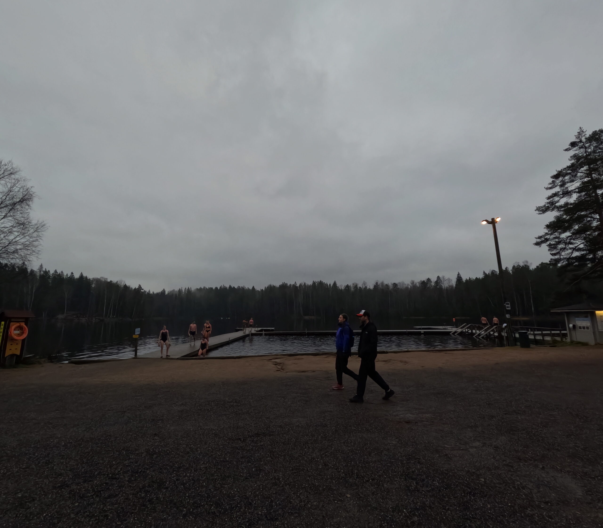 Kuusijärvi湖畔ビーチエリア｜曇り空の下、桟橋から湖に入る複数の水泳者、街灯と砂浜がある冬季水泳スポット｜ヴァンター市・フィンランド
