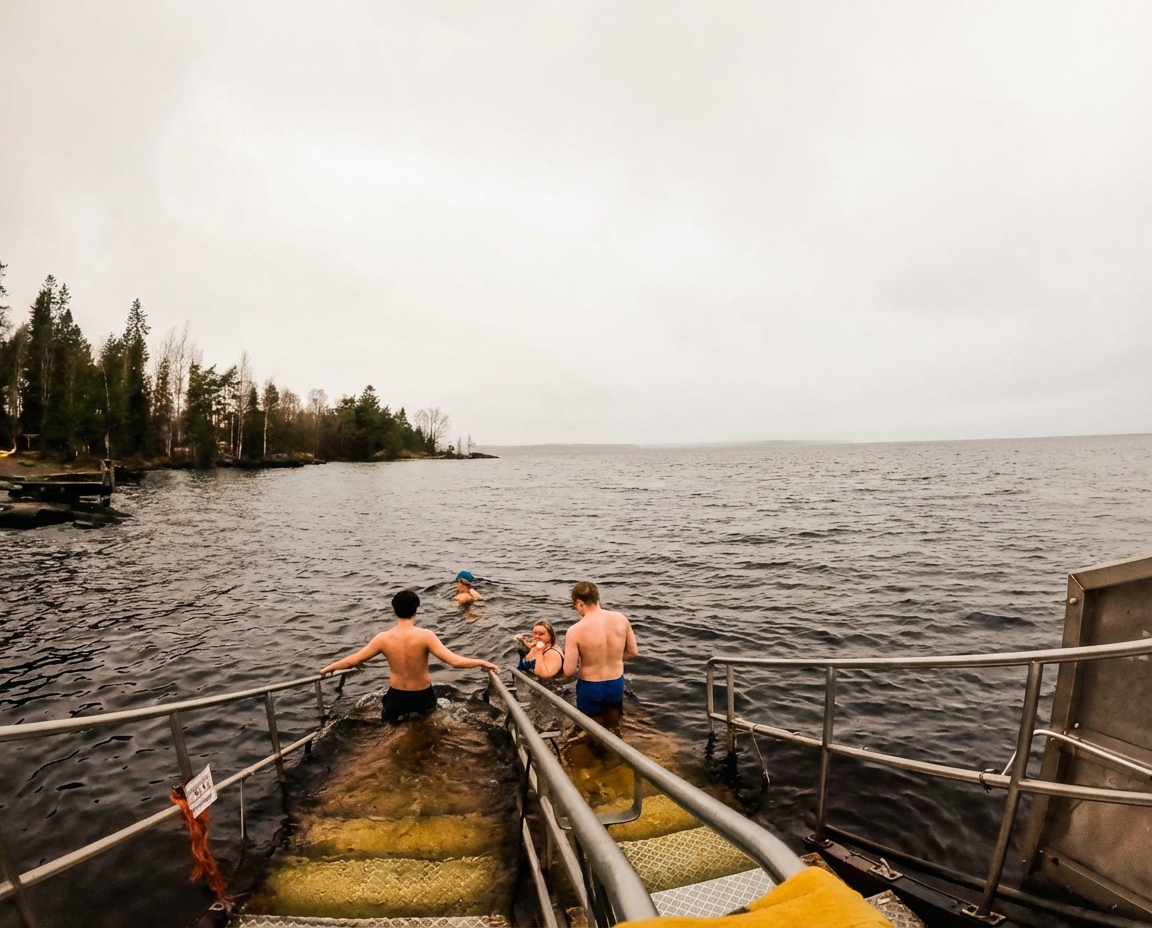 Rauhaniemi Folk Spaのナシ湖水風呂体験｜手すり付き階段から湖に入る水着姿の男女、曇り空の下広がる湖面と対岸の針葉樹林｜体感シングルの天然水風呂｜フィンランド・タンペレ
