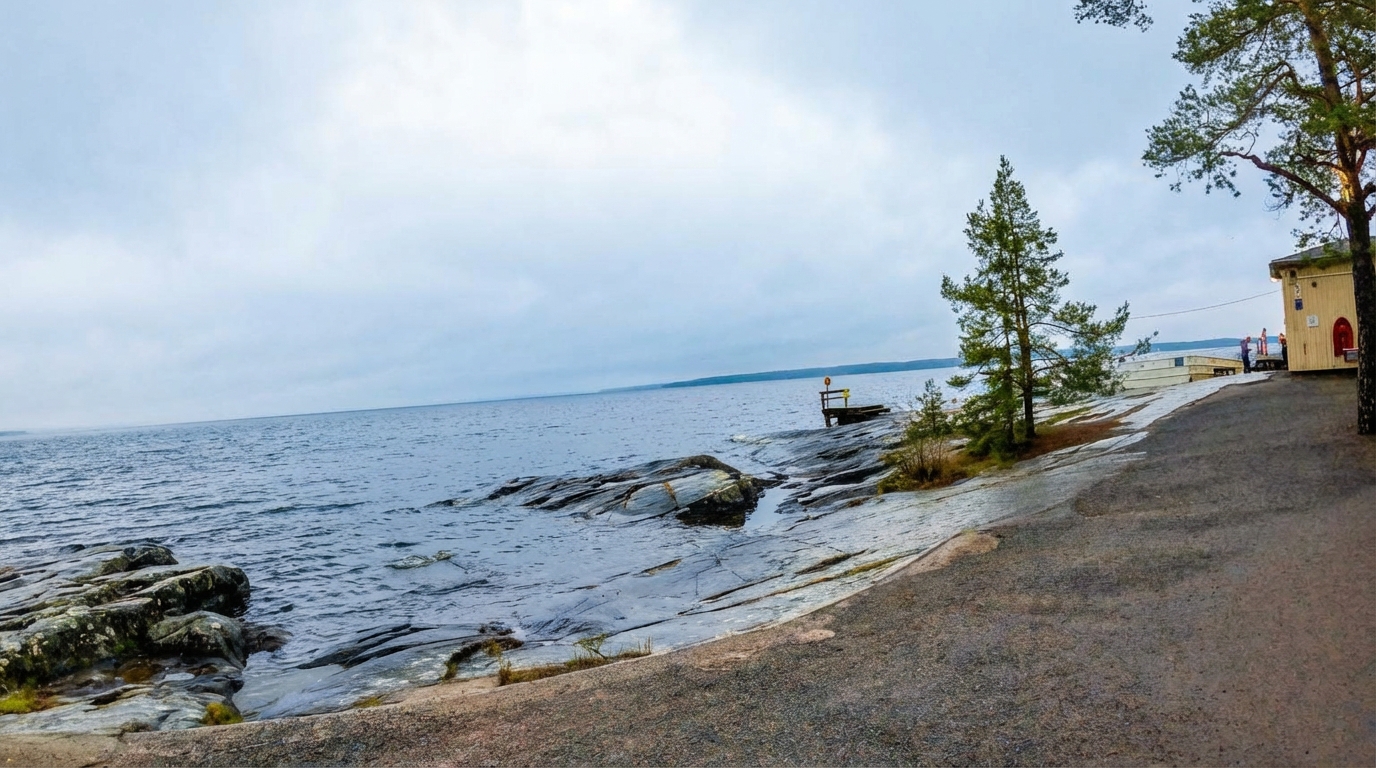 Rauhaniemi Folk Spaの湖畔景観｜右手に黄色いサウナ施設、左手にナシ湖の広大な水面、岩場の湖岸と針葉樹、曇り空の下の静かな北欧の風景｜サウナ後の外気浴に最適な環境｜タンペレ
