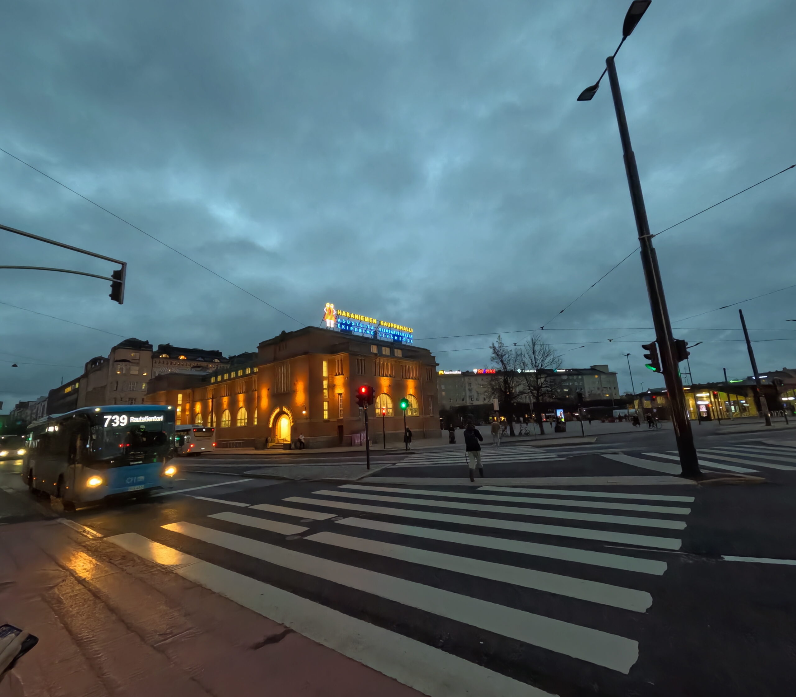 ハカニエミ駅周辺｜オレンジ色にライトアップされた駅舎建物、横断歩道を渡る人々、バスが走る夕暮れの街並み、屋上に「HAKANIEMI」のネオンサイン｜ヘルシンキ中心部