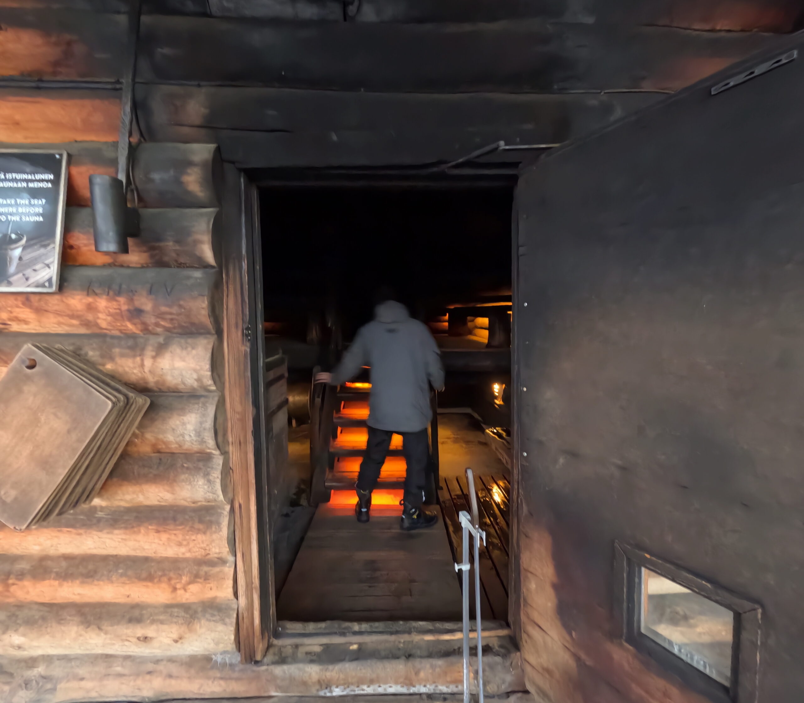 Kiilopääスモークサウナ室内入口｜薄暗いサウナ室に入る人の後ろ姿、オレンジ色に光る階段照明、煤で黒く染まった木製壁面｜フィンランド・サーリセルカ・北極圏