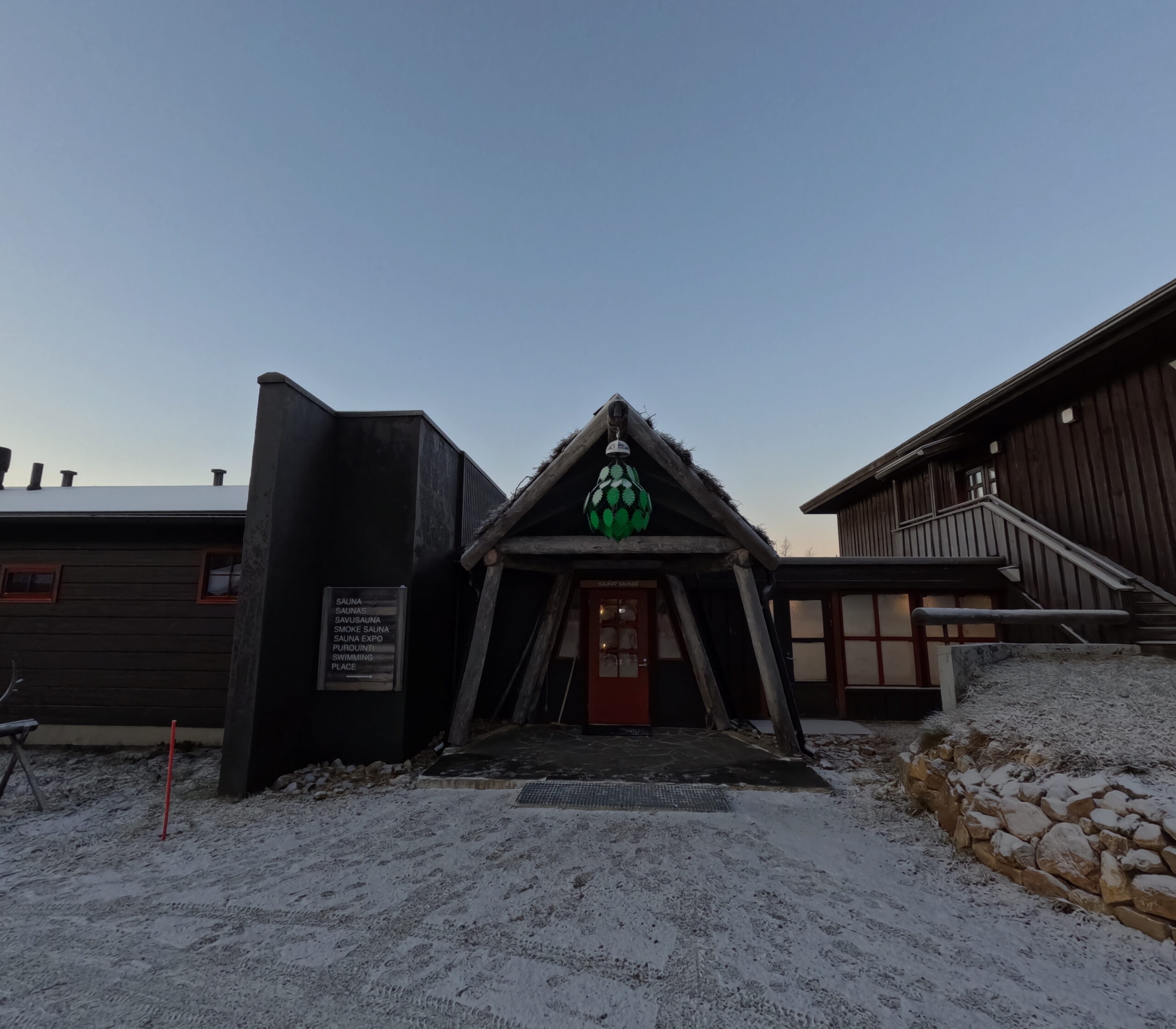 Kiilopää施設外観｜三角形の丸太フレーム入口、緑色の装飾ランプが頂点を飾る、雪が積もった地面、右側に窓から温かい光が漏れる建物｜夕暮れ時｜フィンランド・サーリセルカ・北極圏