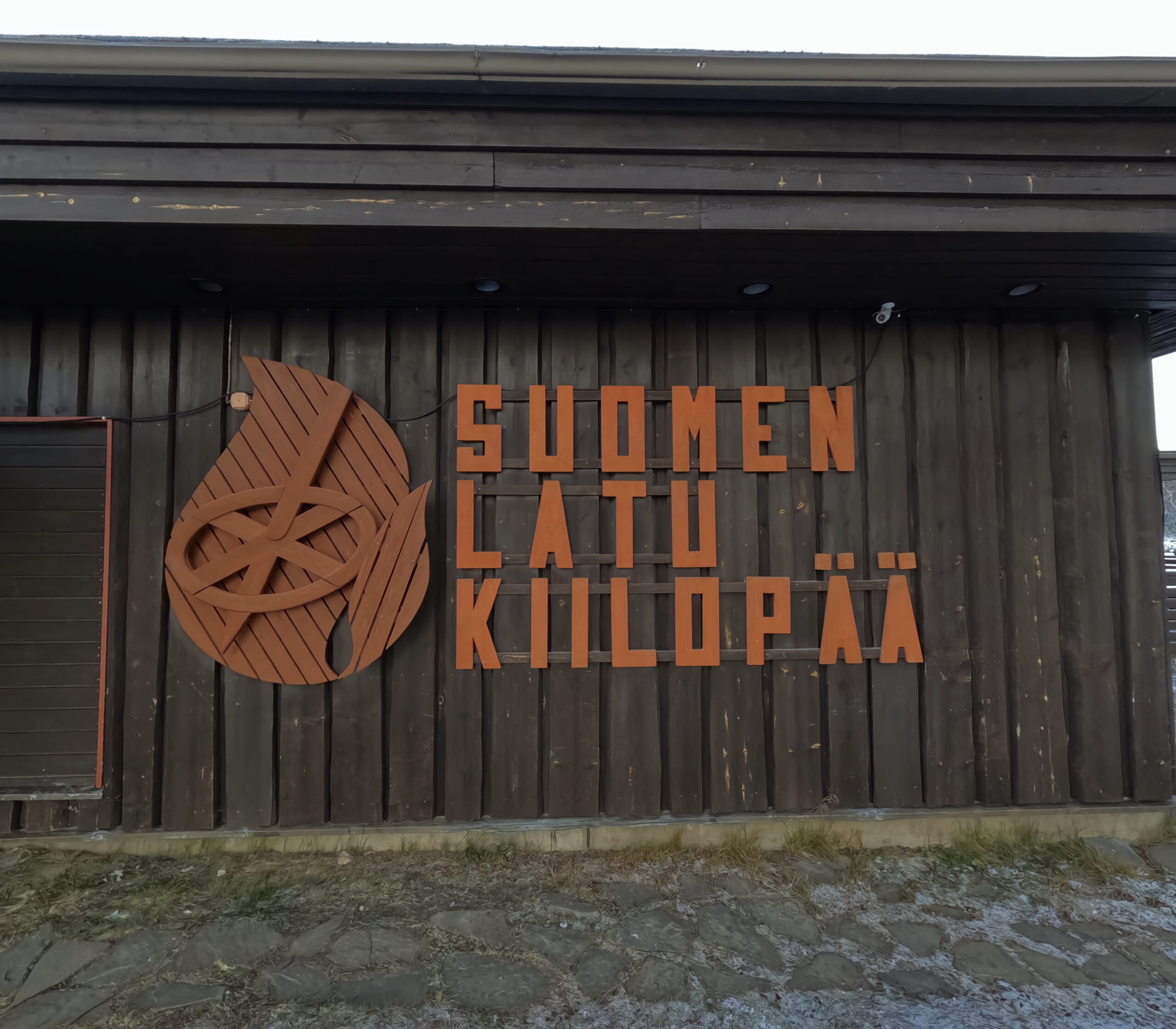 Kiilopää施設看板｜「SUOMEN LATU KIILOPÄÄ」と刻まれた大型木製サイン、オレンジ色の文字と木目のロゴマーク、丸太の壁面｜フィンランド・サーリセルカ