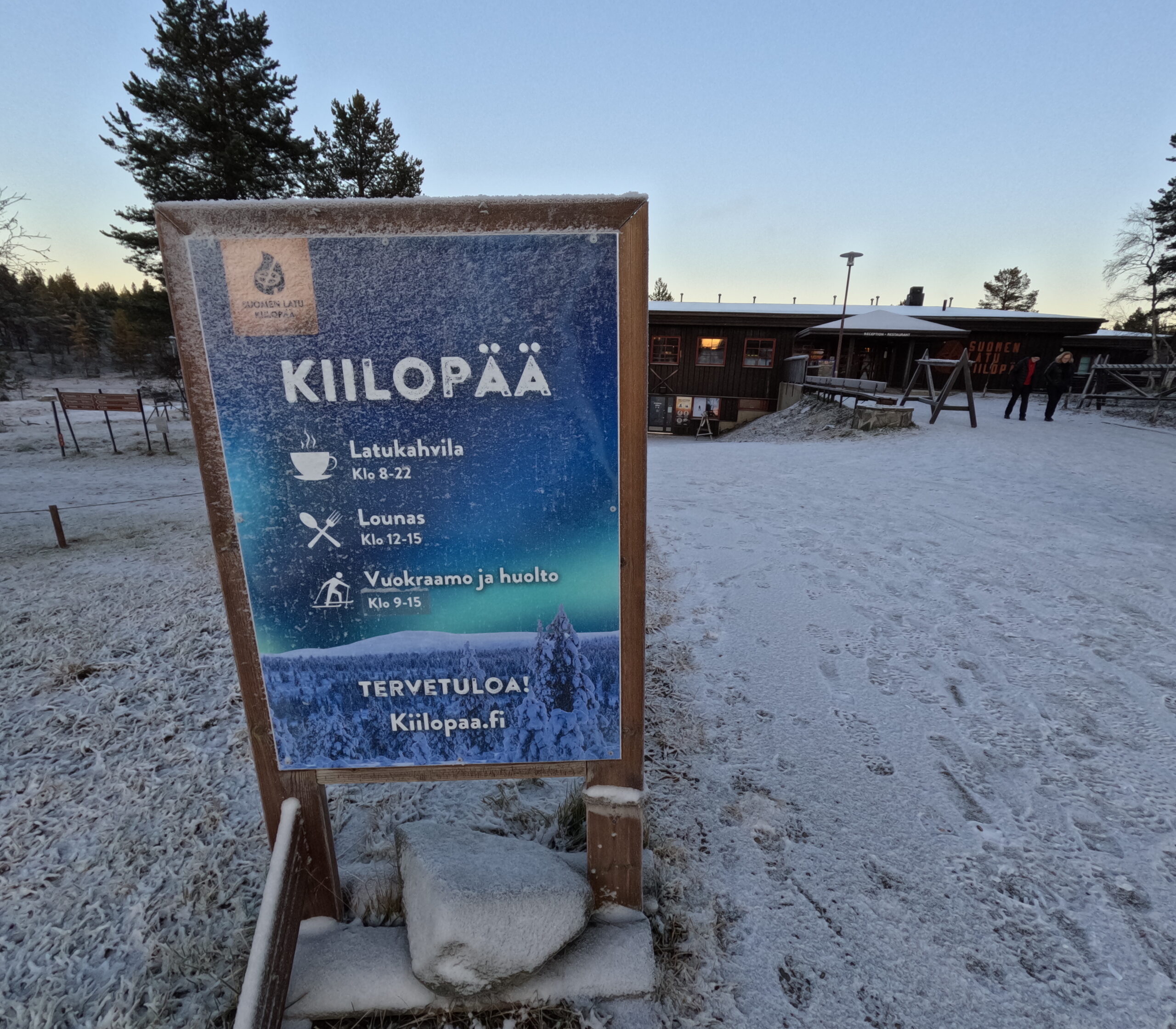 Kiilopää施設案内ボード｜「KIILOPÄÄ」の大型看板、オーロラ写真が背景、カフェ・ランチ・レンタルの営業時間情報、雪に覆われた木製フレーム｜フィンランド・サーリセルカ