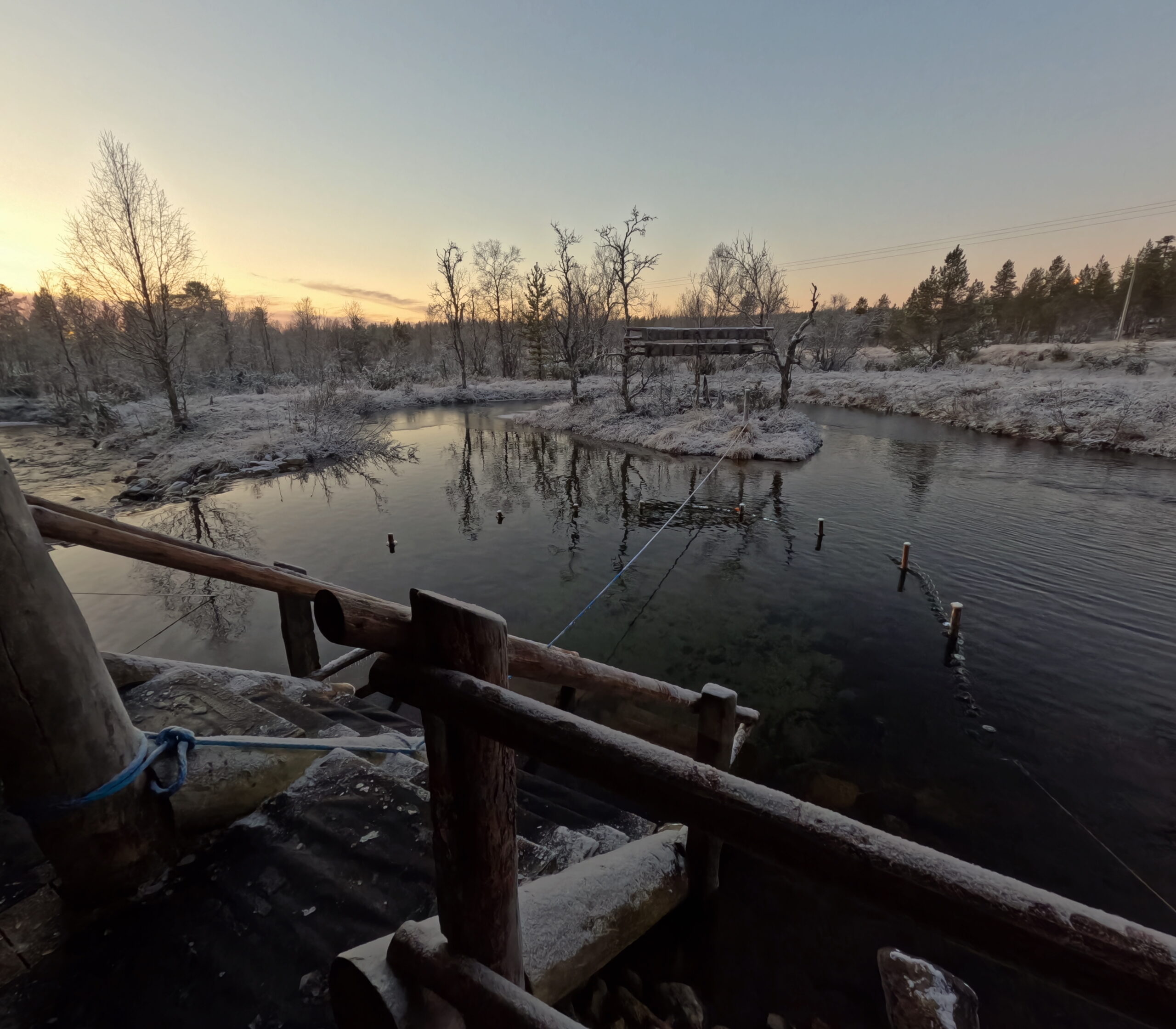 Kiilopää小川の水風呂全景｜夕焼け空の下、木製デッキと手すりから続く階段、霜で白く染まった岸辺と針葉樹林、静かに流れる清流｜フィンランド・サーリセルカ・北極圏