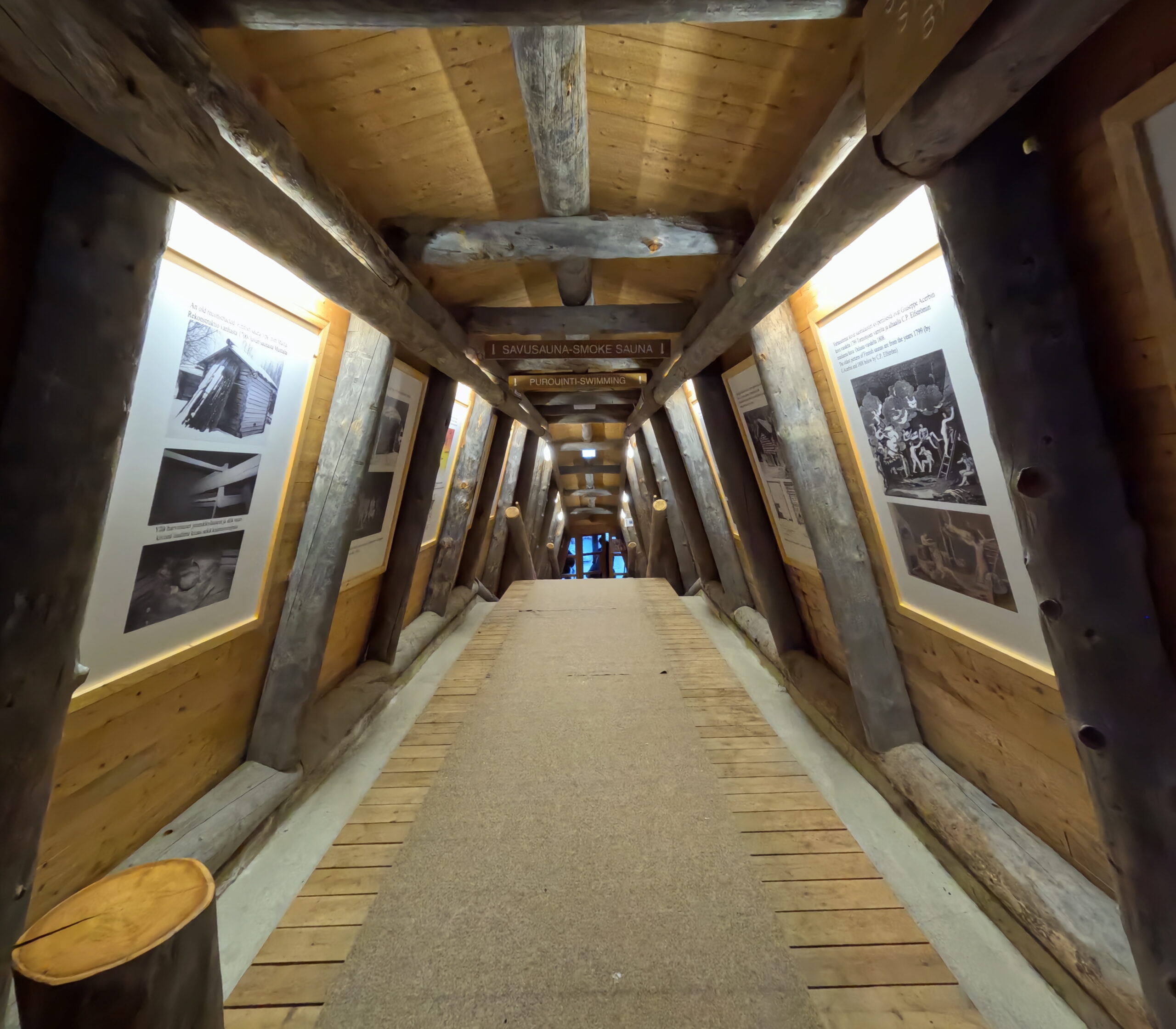Kiilopääサウナへ続く通路｜丸太造りの長い廊下、壁面に「SAVUSAUNA SMOKE SAUNA」の案内板と歴史写真の展示、間接照明で照らされた温かい雰囲気｜フィンランド・サーリセルカ・北極圏