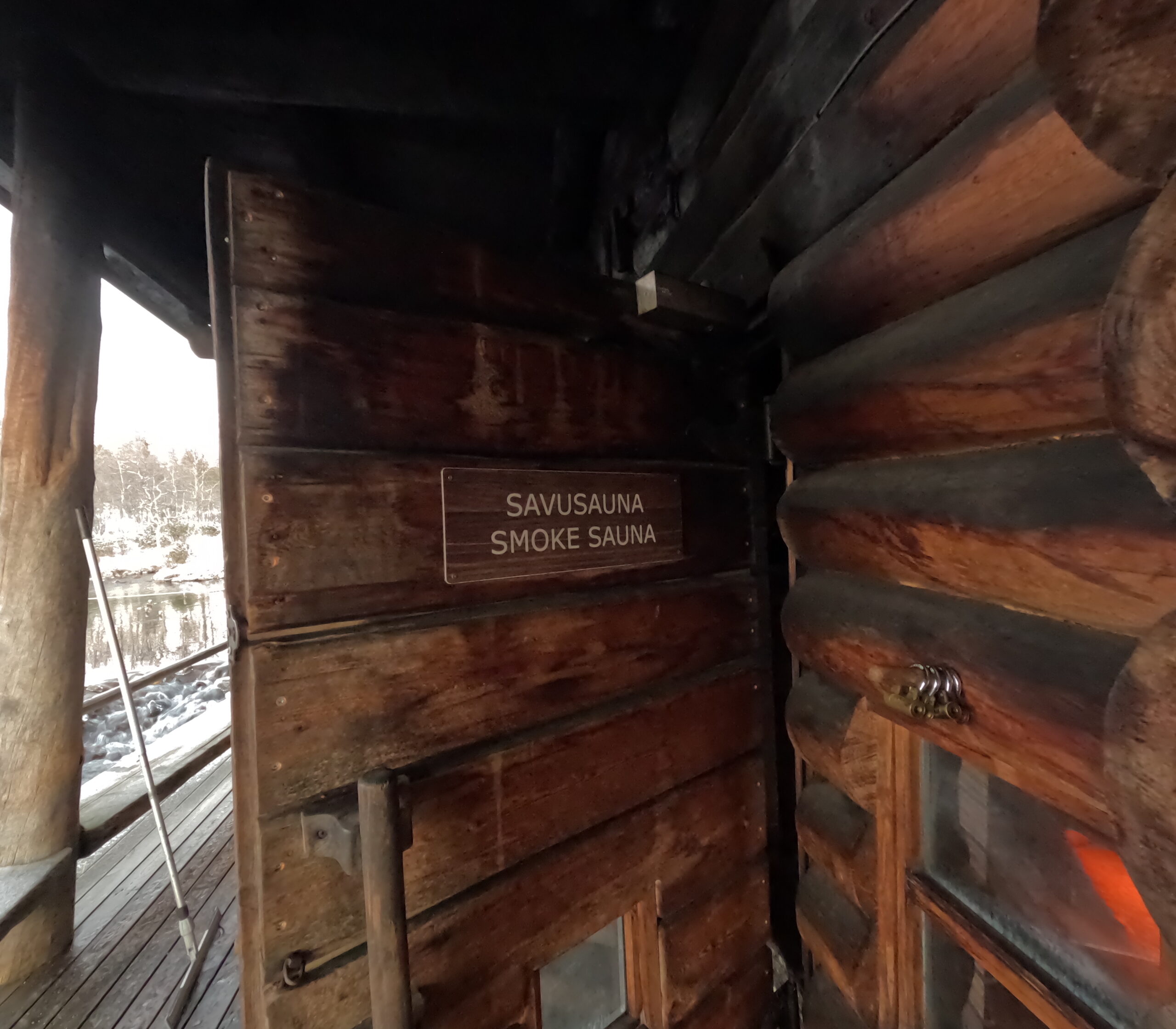 Kiilopääスモークサウナ扉｜煤で黒く染まった丸太壁、「SAVUSAUNA SMOKE SAUNA」の木製看板、ガラス窓から赤く光るストーブが見える重厚な入口｜フィンランド・サーリセルカ・北極圏
