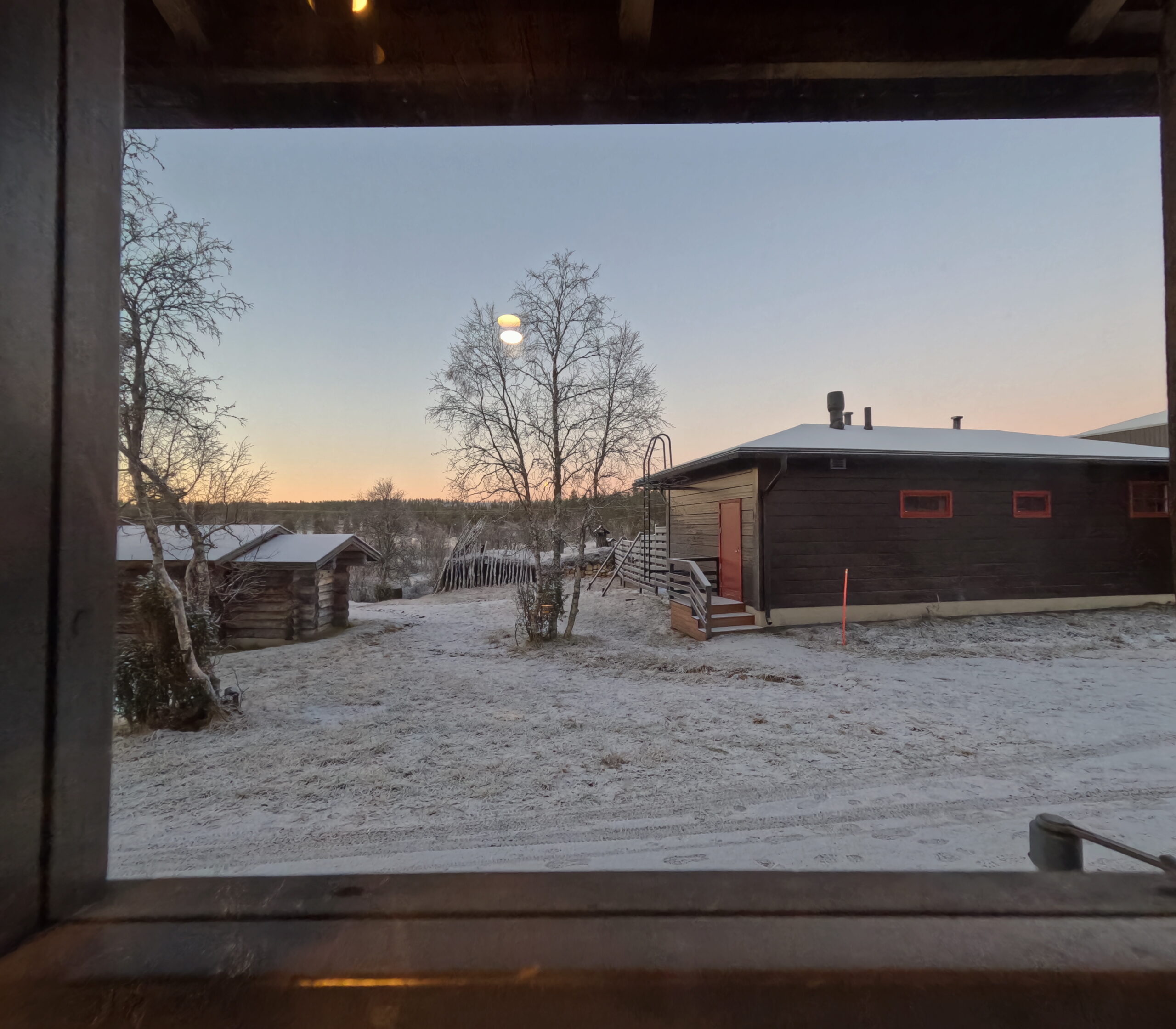 Kiilopääの宿泊施設から窓越しに眺める冬の雪景色｜木々と木製建物が点在する静謐な夕暮れの風景｜フィンランド・ラップランド地方