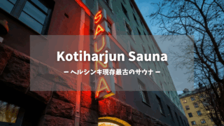 Kotiharjun Saunaアイキャッチ画像｜夕暮れに輝く赤いネオン縦看板「SAUNA」、石造り建物外壁、グレーとオレンジのアパート、街路樹、「ヘルシンキ現存最古のサウナ」テキスト入り｜1928年創業の伝統公衆サウナ｜ヘルシンキ・フィンランド