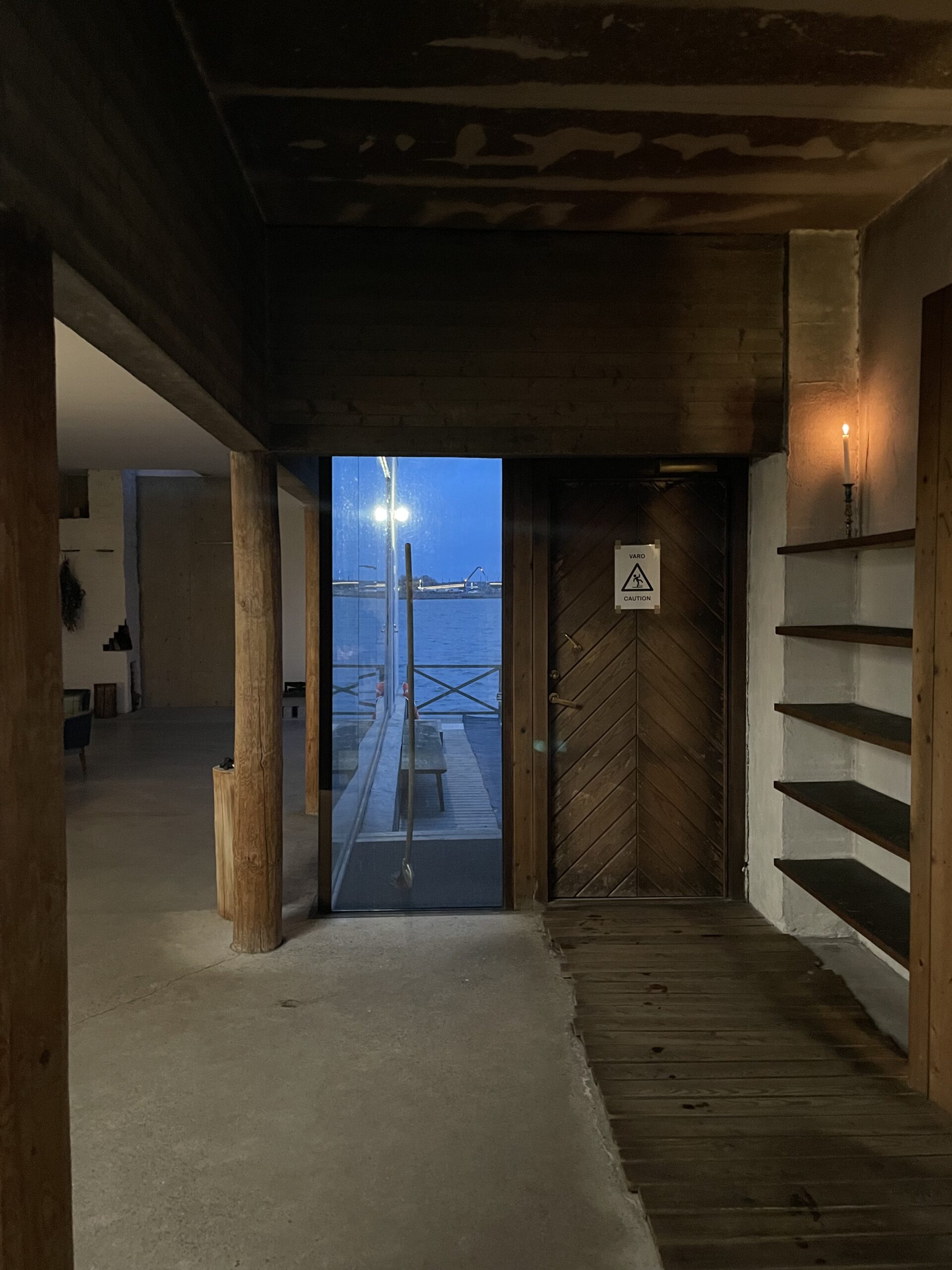 Kulttuurisauna内部エントランス｜コンクリート床と木製の柱が融合した薄暗い空間、奥にバルト海が見える大きな窓、右手にサウナ室の木製ドア、左手にロッカーエリア｜ヘルシンキ