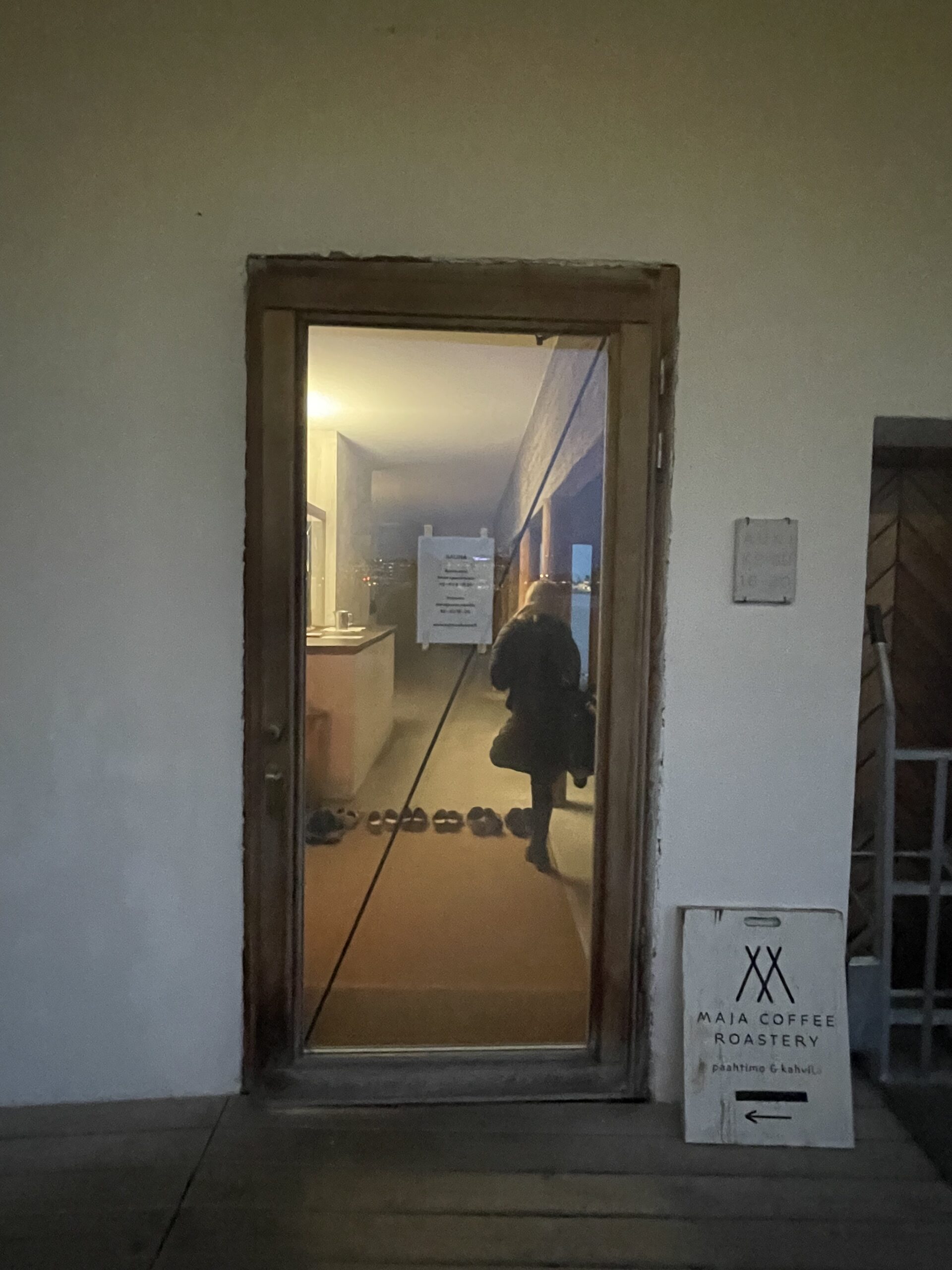 Kulttuurisauna入口｜コンクリート壁の開口部から見えるおしゃれな内装、靴が並ぶ木製床、右手にMAIA COFFEE ROASTERYの看板、左手にシューズラック｜ヘルシンキ