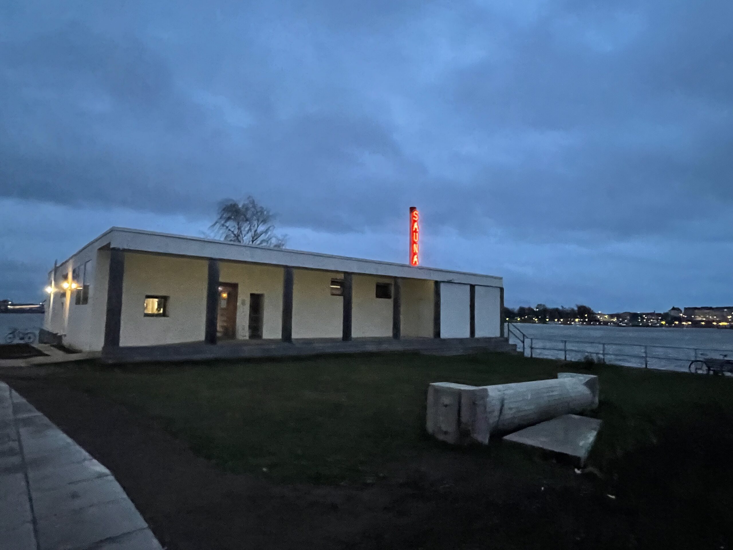 Kulttuurisauna外観｜バルト海沿いに佇む白い平屋建てのサウナ施設、屋上に赤いネオンサイン「SAUNA」が灯る夕暮れの景色、手前に芝生と流木ベンチ、奥にヘルシンキの街並み｜フィンランド・ヘルシンキ