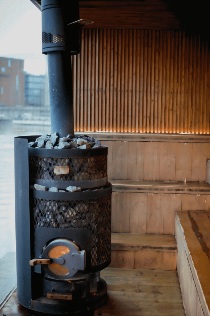 Kuuma屋内薪サウナ室｜黒い円柱型ikiストーブと大量のサウナストーン、木製3段座面、大きな窓から川の景色、間接照明の温かい雰囲気｜フィンランド・タンペレ