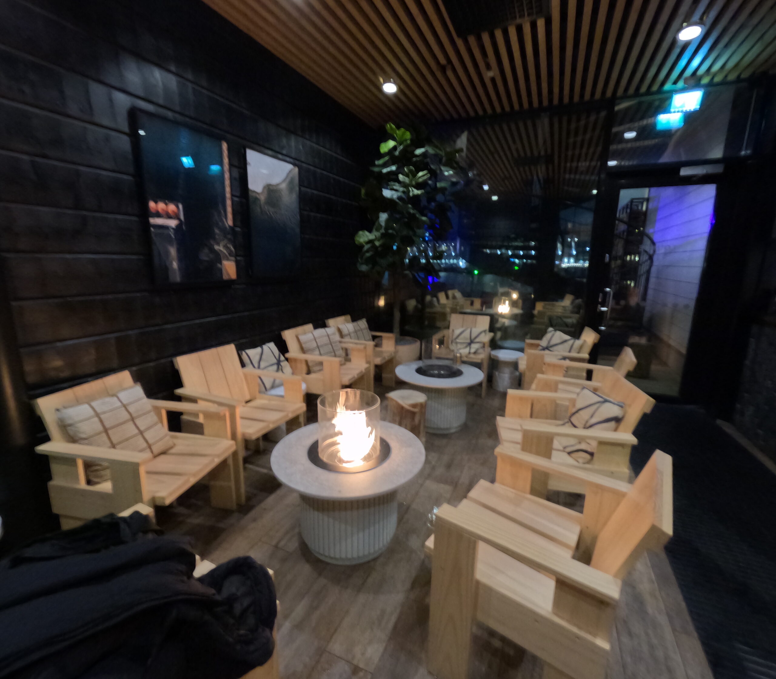 Kuuma休憩ラウンジ｜黒を基調としたシックな空間、木製ソファと丸テーブル、暖炉、観葉植物、間接照明、川の夜景が見える大きな窓｜フィンランド・タンペレ