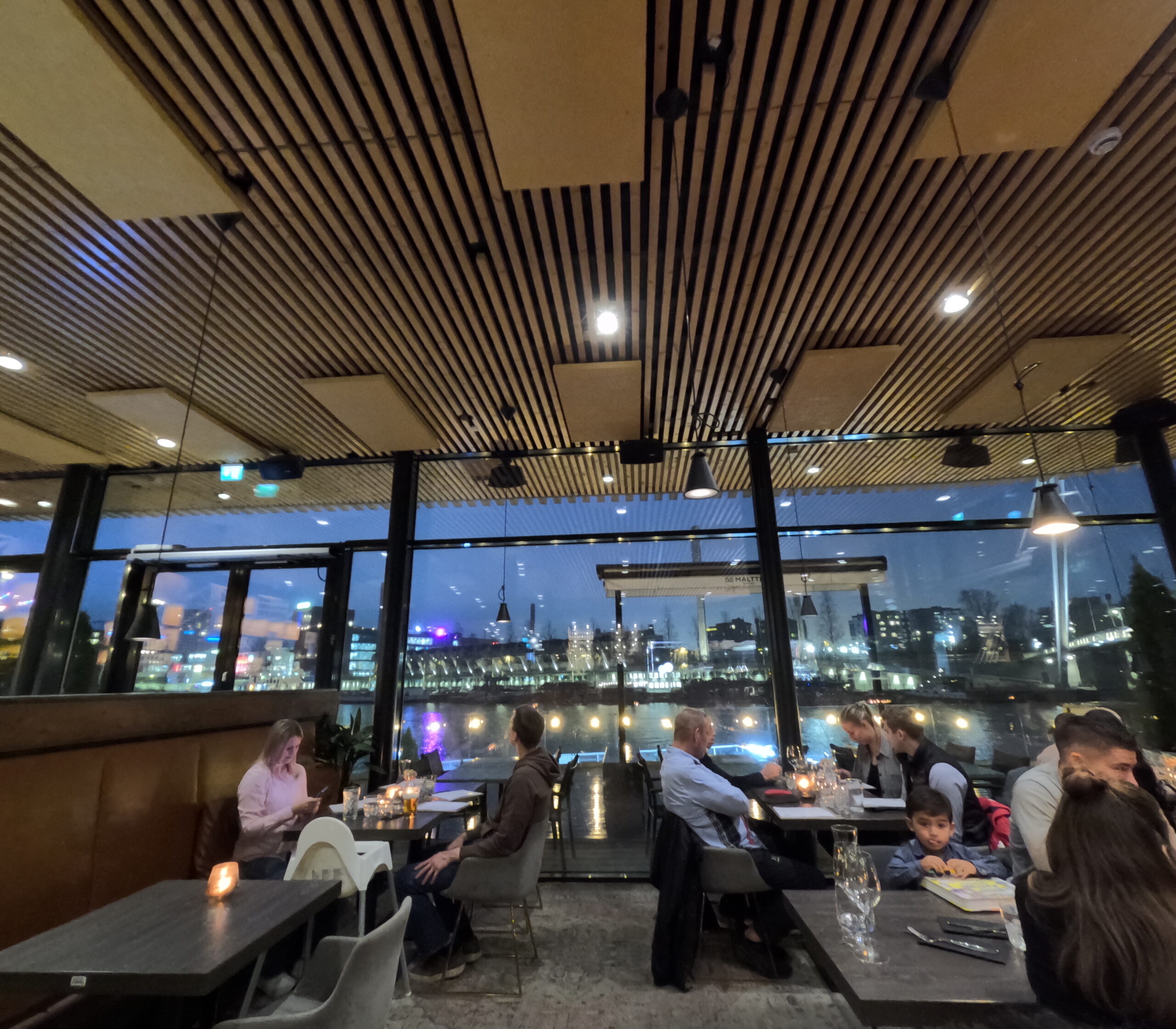Kuumaレストラン店内｜木製ルーバー天井、大きな窓から川と市街地夜景、テーブル席で食事する人々、温かい照明の北欧インテリア｜フィンランド・タンペレ