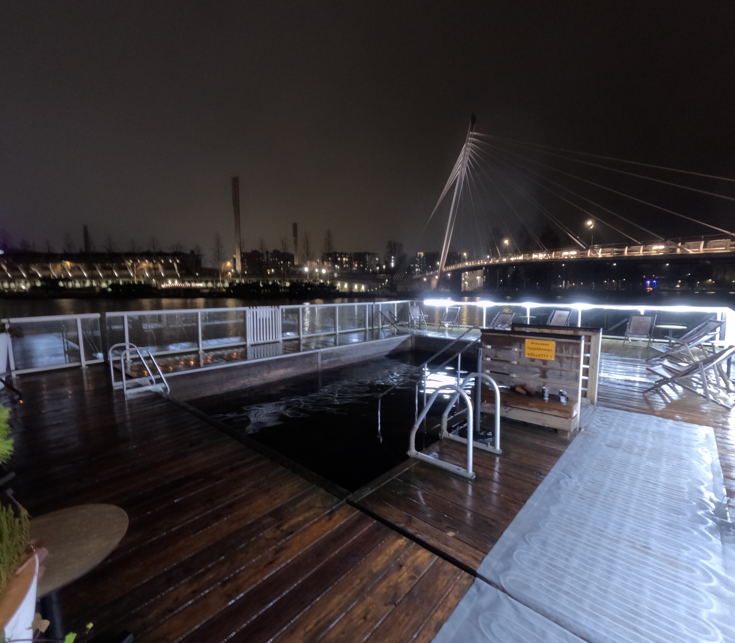 Kuumaウッドデッキと川水風呂プール｜手すり付き桟橋、タンメルコスキ川に設置された水風呂プール、対岸にタンペレ市街地、夜の照明が映る川面｜フィンランド・タンペレ