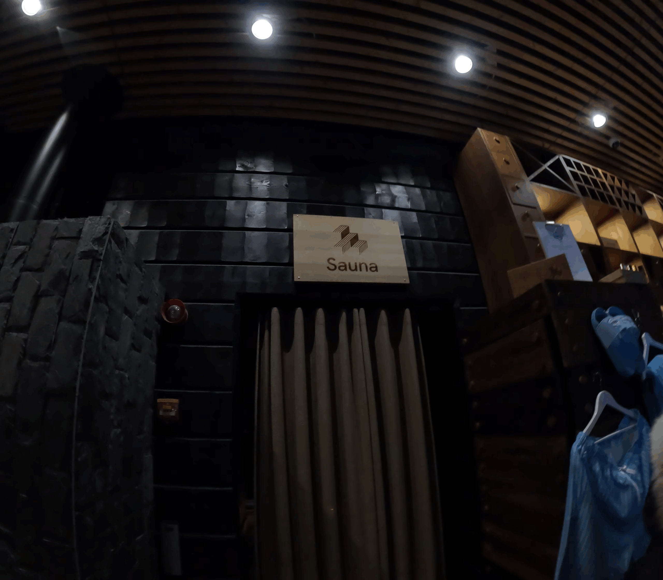 Kuumaサウナ入口｜黒いタイル壁面に「Sauna」白いサイン、木製ルーバー天井、間接照明、更衣室への通路、モダンなデザイン｜フィンランド・タンペレ