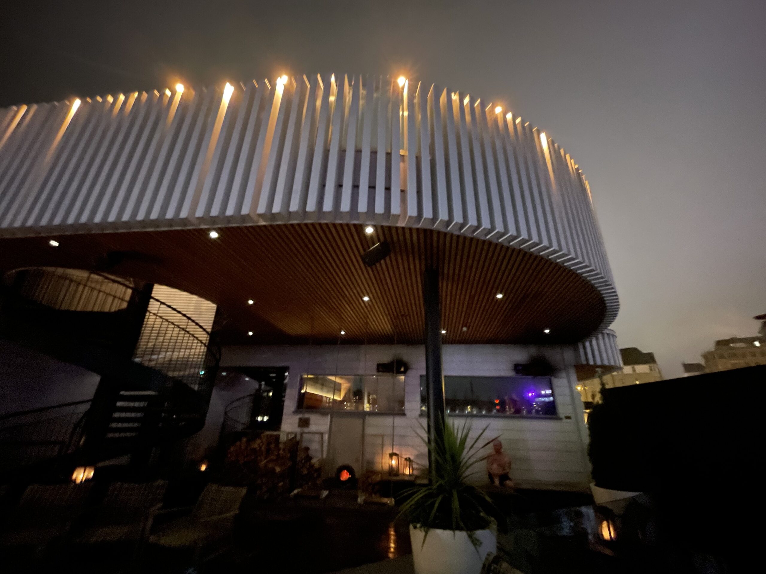 Kuuma建物外観｜白い波型屋根に上部照明、2階レストランテラス、1階エントランス、川沿いの現代建築デザイン、夕暮れの曇り空｜フィンランド・タンペレ