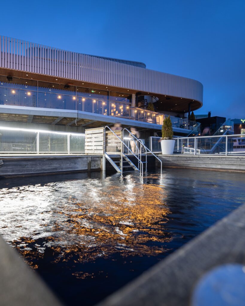 Kuuma（クーマ）川沿い外観とタンメルコスキ川水風呂プール｜白い波型屋根の現代建築、ライトアップされたレストランテラス、手すり付き桟橋と川水風呂、夕暮れの青空｜フィンランド・タンペレ
