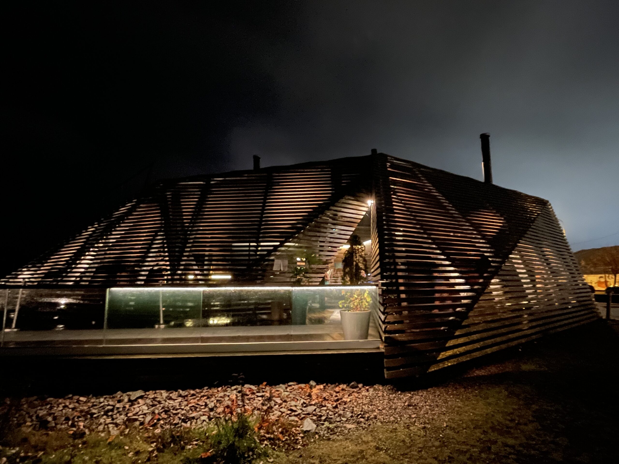 löylyヘルシンキの夜の外観｜波のような曲線を描く木製ルーバーの建築美、地面に埋め込まれたガラス窓から漏れる光、手前には岩場が広がる現代的デザイン｜フィンランド・ヘルシンキ湾岸