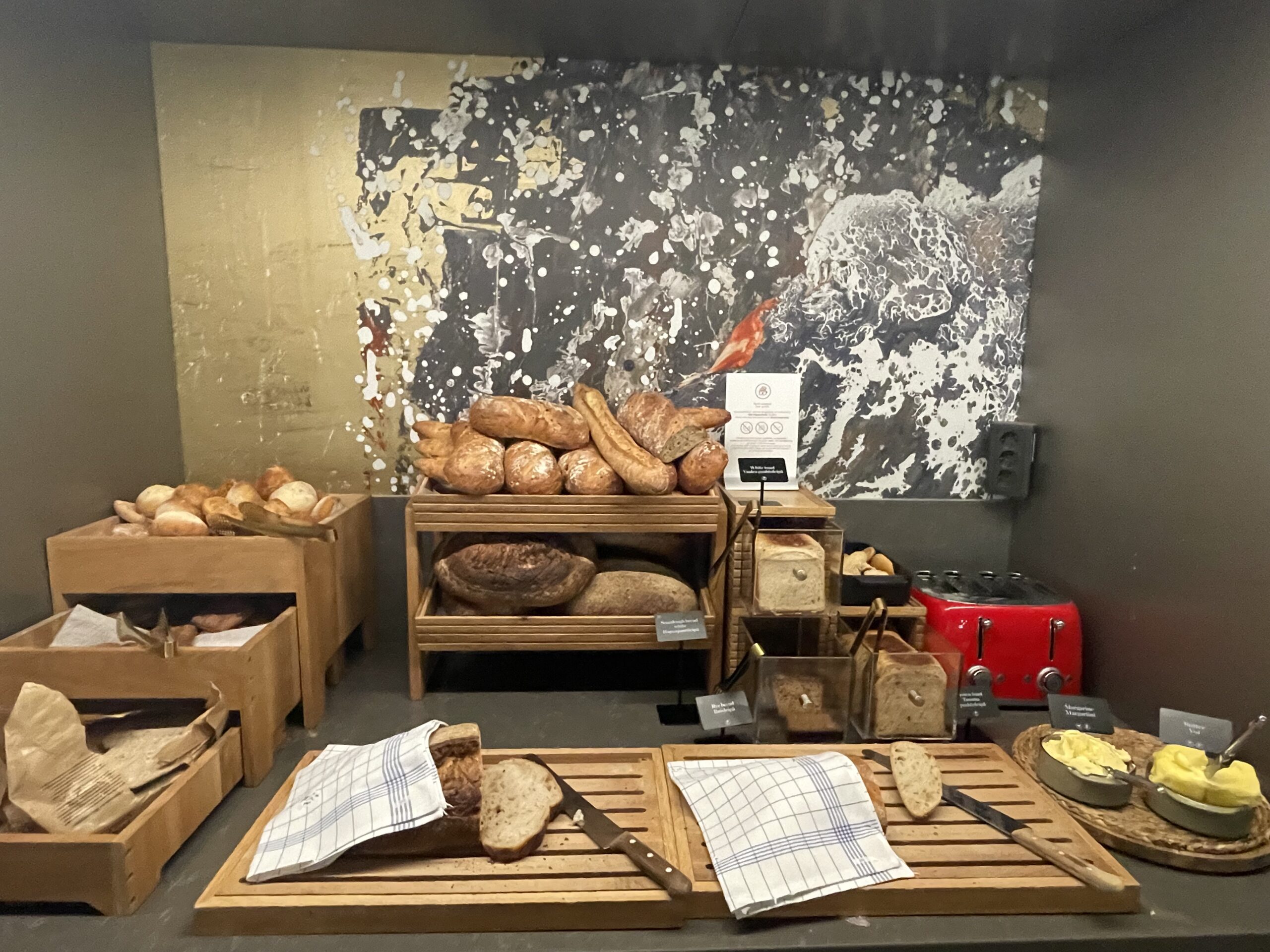 NH Collection Helsinki Grand Hansa朝食ビュッフェのブレッドコーナー｜バゲット・職人技の焼きたてパンが木製トレイに並ぶ、ダークグレーの背景が素材の美しさを引き立てる｜ヘルシンキ・フィンランド