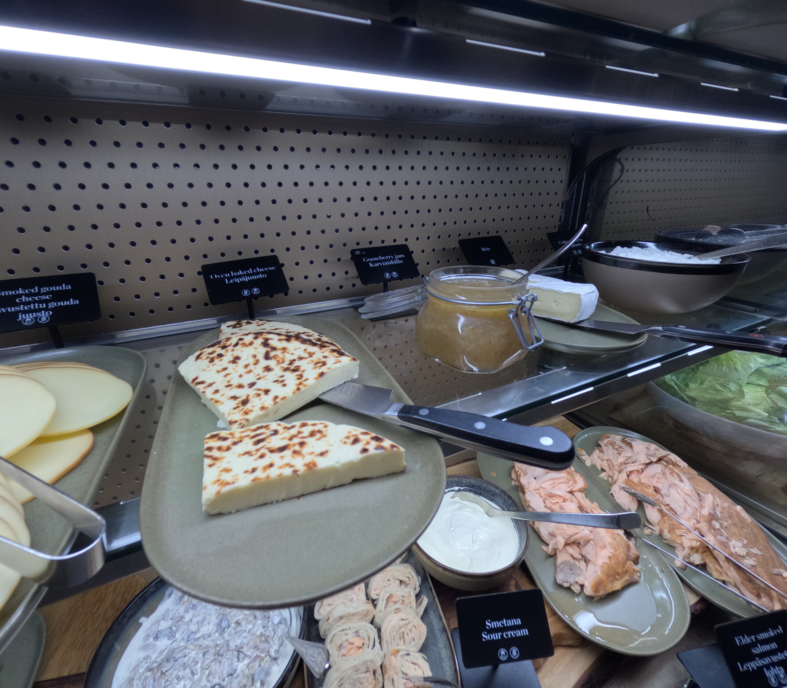 NH Collection Helsinki Grand Hansa朝食ビュッフェのチーズ・魚コーナー｜焼きチーズ・スモークサーモン・サワークリーム・クランベリージャムが冷蔵ショーケースに並ぶ、明るい照明で新鮮さを強調｜ヘルシンキ・フィンランド