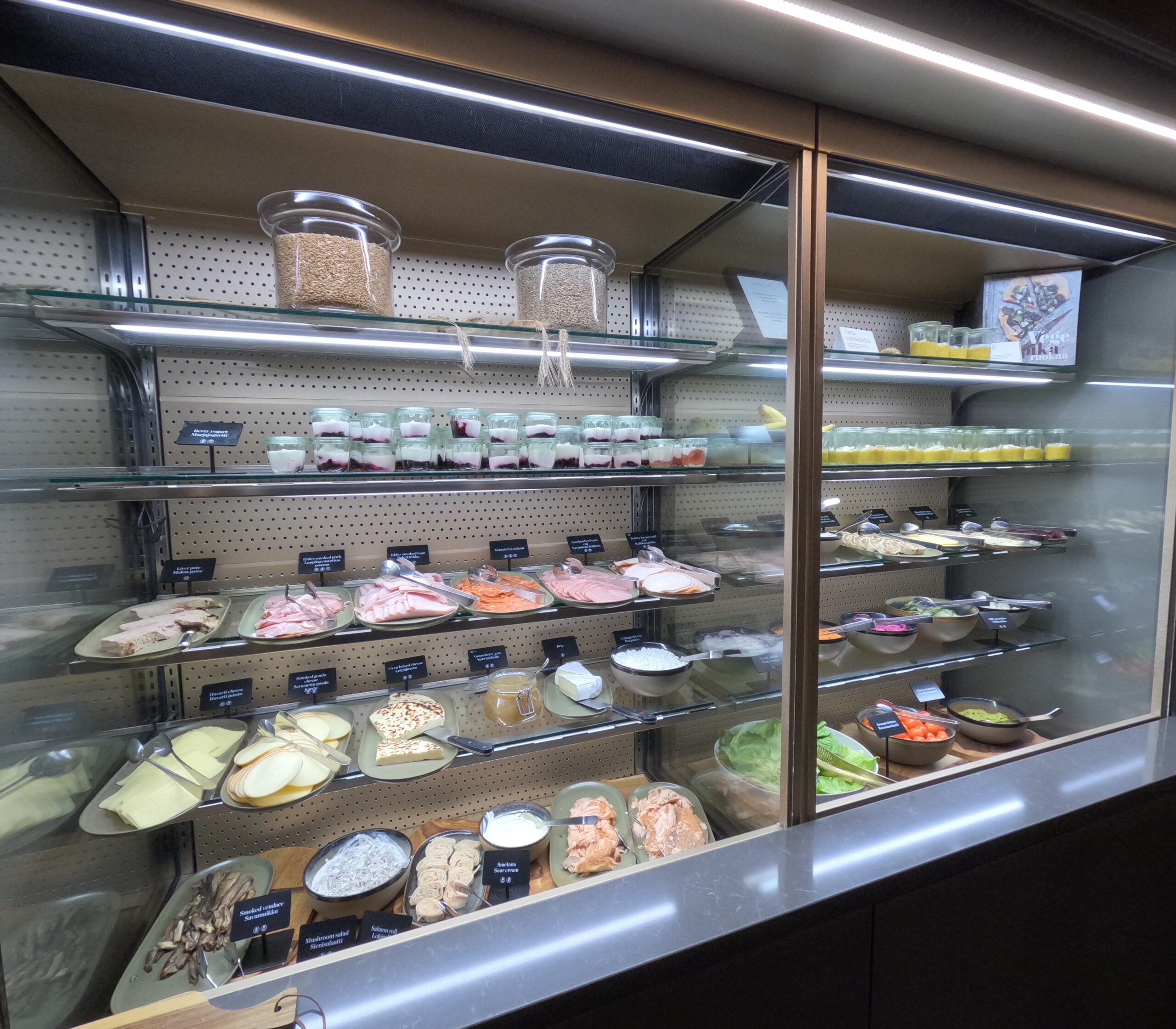 NH Collection Helsinki Grand Hansa朝食ビュッフェの冷蔵セクション全景｜チーズ・ハム・スモークフィッシュ・ヨーグルト・プディングが整然と並ぶガラス棚、メタルラックと明るい照明で豪華さを演出｜ヘルシンキ中央駅前・フィンランド