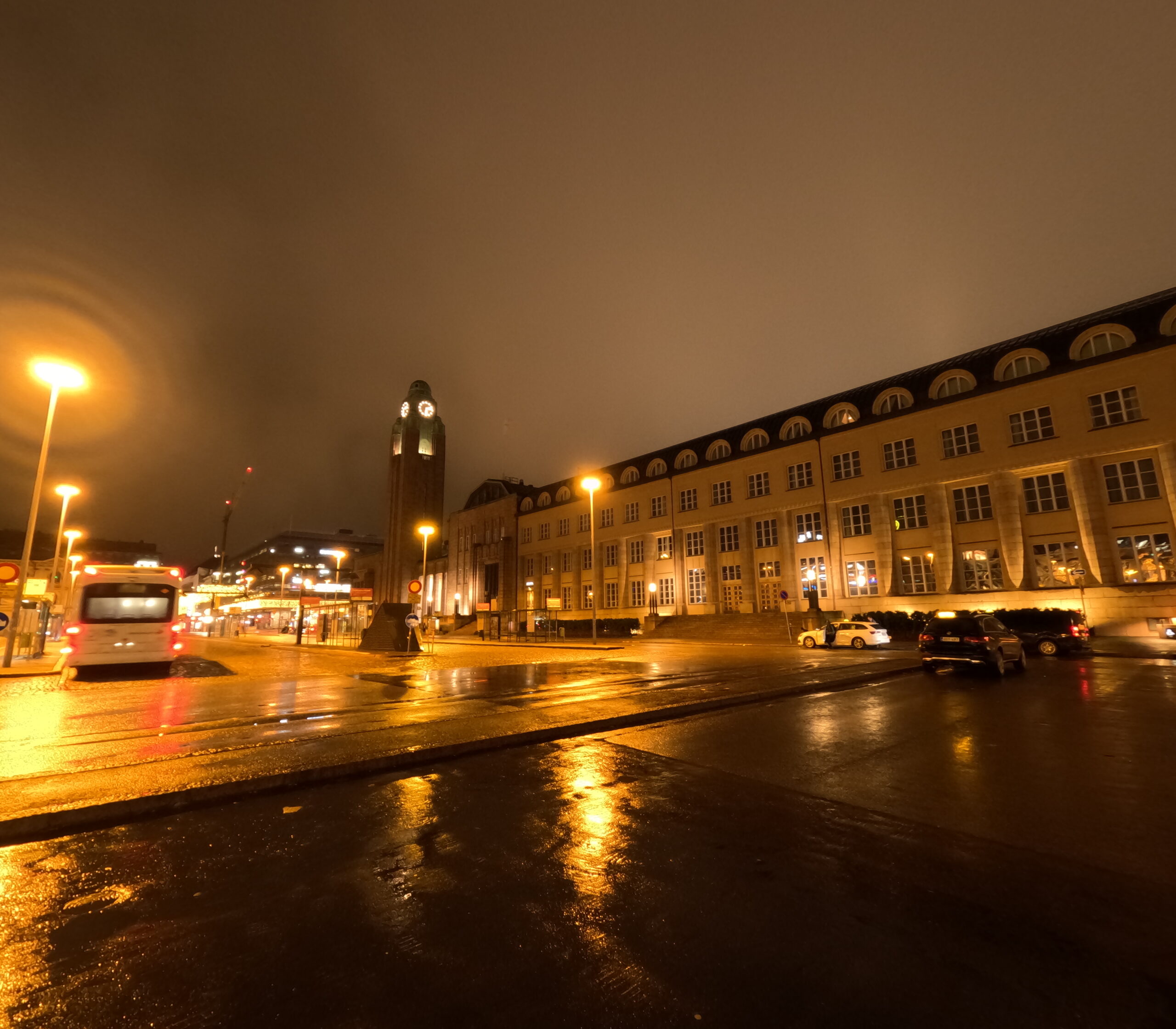 Scandic Grand Central Helsinki夜の外観｜雨に濡れたロータリー、ライトアップされたクリーム色のホテル建築、奥にヘルシンキ中央駅の緑の時計塔、バスとタクシーが行き交う夜景｜フィンランド・ヘルシンキ