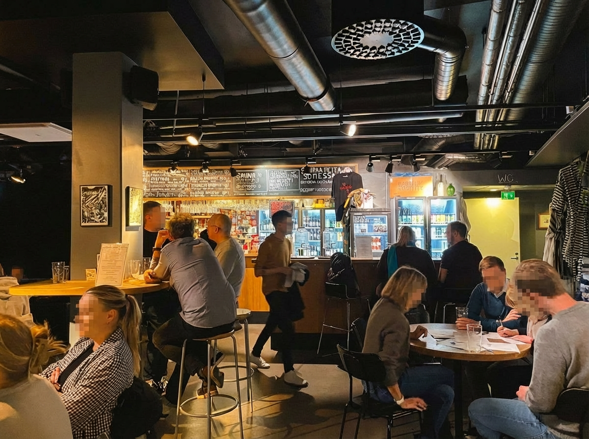 uusi saunaバー・レストランエリア｜黒い天井と木製カウンター、冷蔵ケースにフィンランドビールが並ぶバーカウンター、テーブル席で食事を楽しむ人々、インダストリアルデザインの照明｜ヘルシンキ