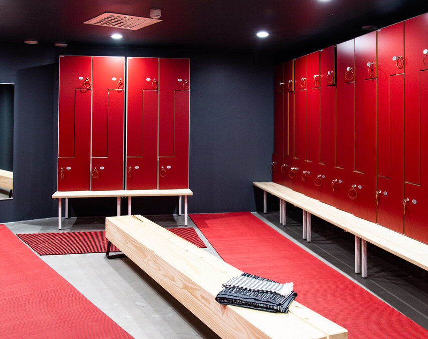 uusi sauna男性用ロッカールーム｜鮮やかな赤色のロッカー扉が両壁に並ぶ、黒い天井と木製ベンチ、赤いマット、海外サッカー風のスタイリッシュな更衣室｜ヘルシンキ都市型サウナ