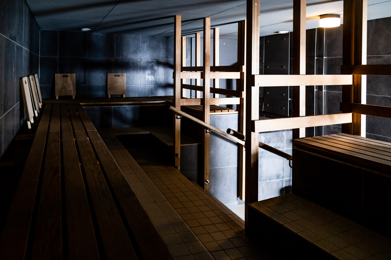 uusi sauna男性用薪サウナ室内｜入口正面に巨大な薪ストーブ、黒タイル壁と木製3段コの字型座面、サウナマット6枚が並ぶ薄暗い照明の伝統的フィンランドサウナ空間｜ヘルシンキ