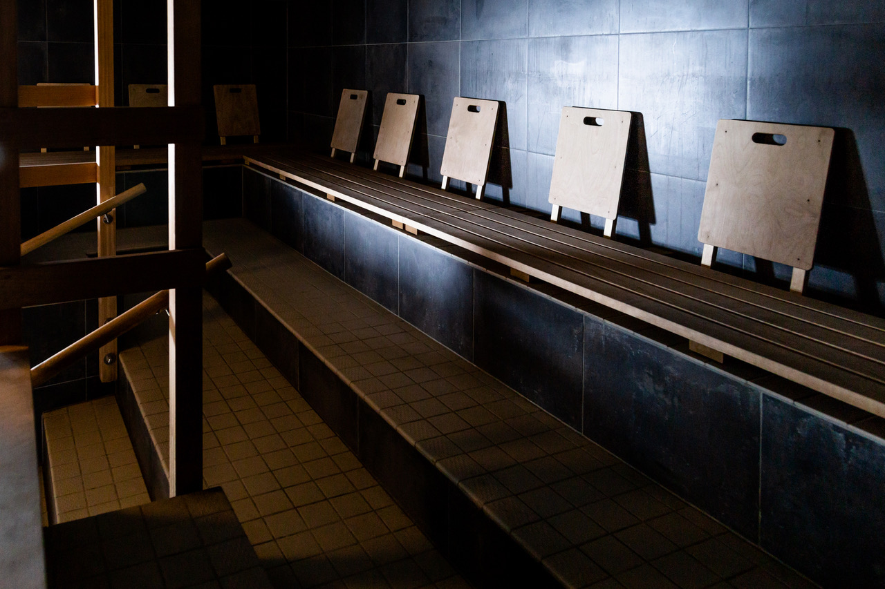 uusi saunaサウナ室内座面詳細｜黒タイル壁に木製サウナマット5枚が立てかけられた3段座面、薄暗い照明とスポットライトで照らされるシンプルで美しい空間｜フィンランド・ヘルシンキ