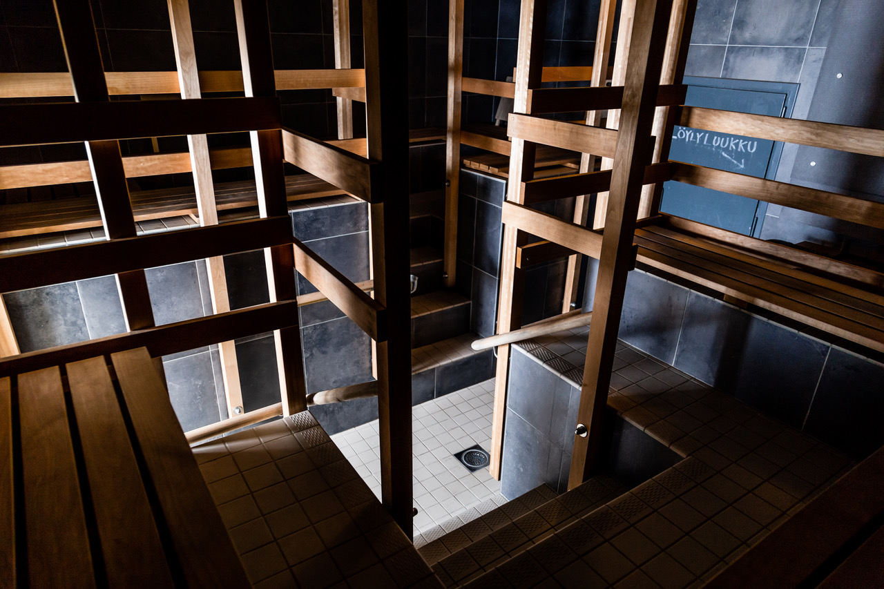 uusi sauna男性用サウナ室階段部分｜木製階段の上から見下ろすシャワールーム、左右にコの字型座面、黒タイル壁と木製ベンチの組み合わせが美しいフィンランド薪サウナ｜ヘルシンキ