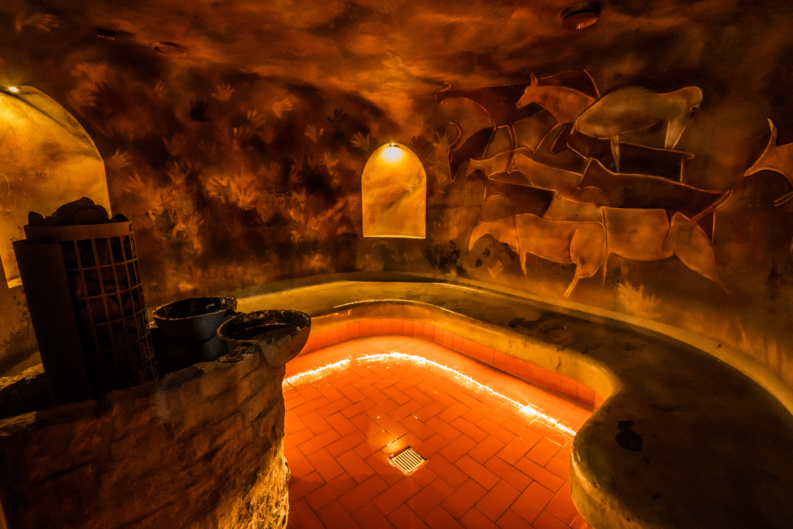 Elamus Spaの泥サウナThe mad hall｜オレンジ色の照明で照らされた円形プール、壁面にアート作品、左側にサウナストーブと木製バケツ、レンガ造りの雰囲気｜全身泥パック体験ができる美容サウナ｜エストニア・タリン