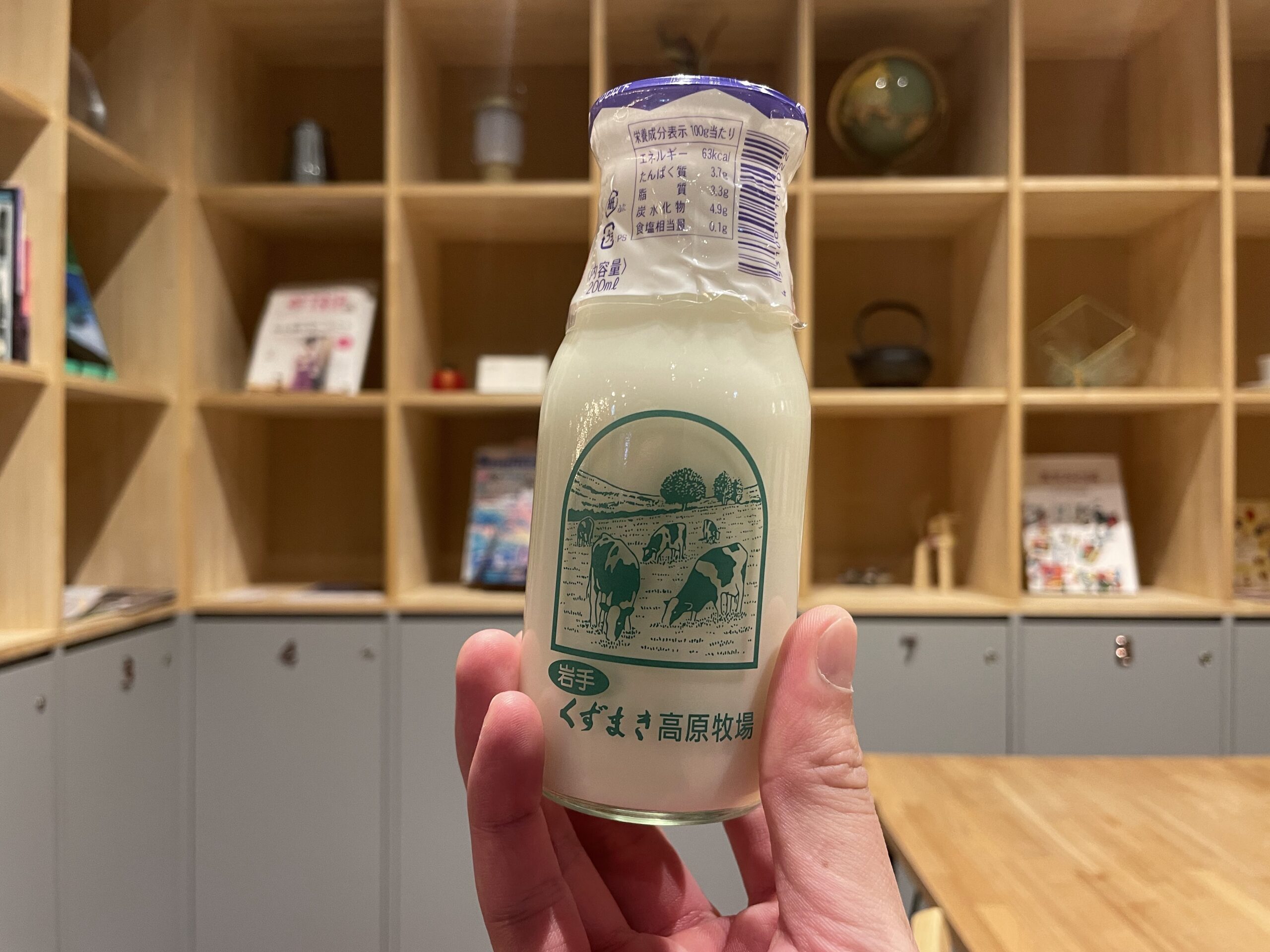 KANAN SPAの湯上がりドリンク｜くずまき高原牧場の牛乳瓶を手にした館内カット、木製シェルフと柔らかな照明が映える休憩空間｜岩手県盛岡市