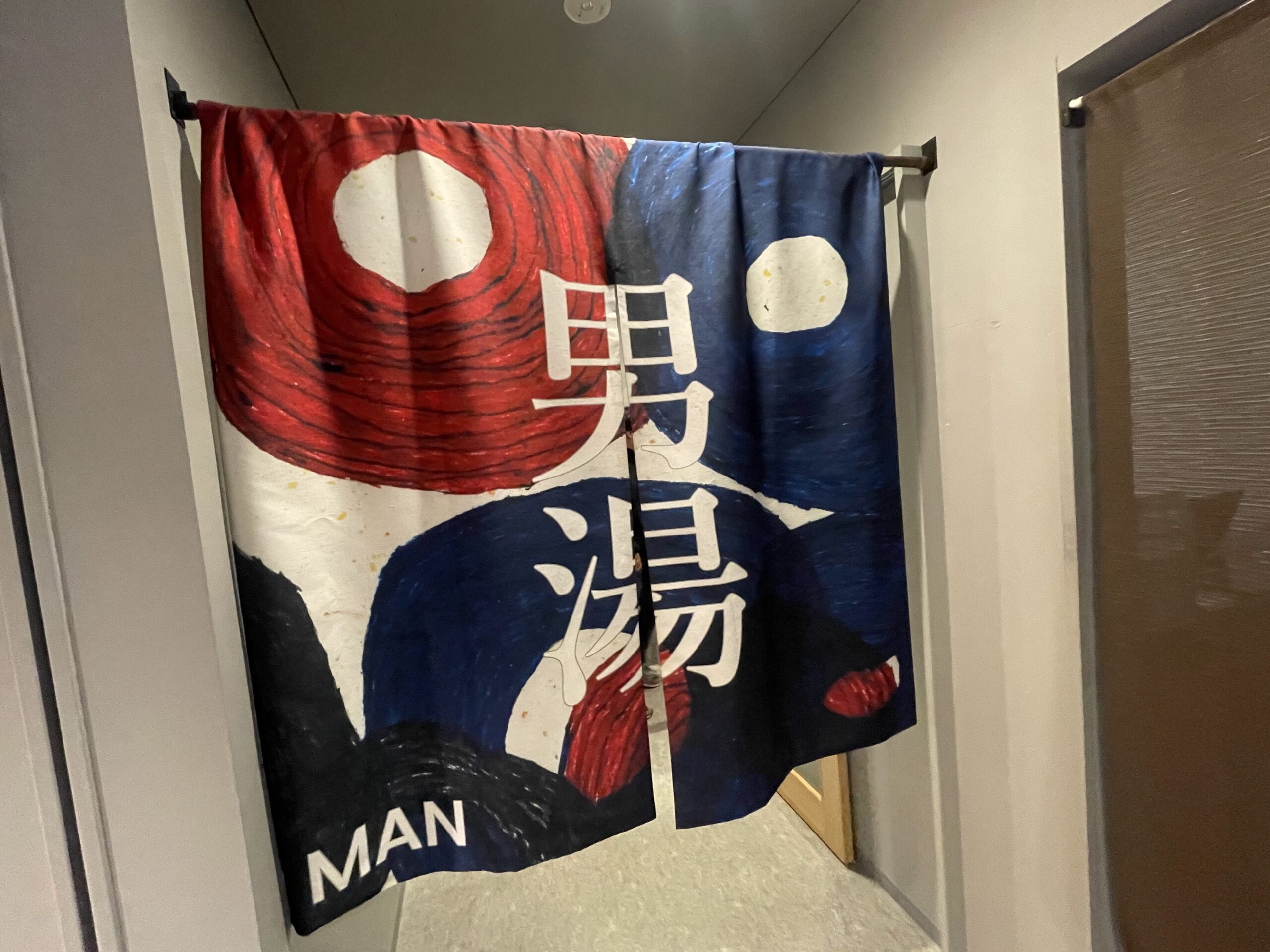 KANAN SPAの男湯入口暖簾｜赤と紺の抽象柄に白字で男湯とMAN表記が入るアート暖簾、木目の扉前に掛かる館内導線｜岩手県盛岡市