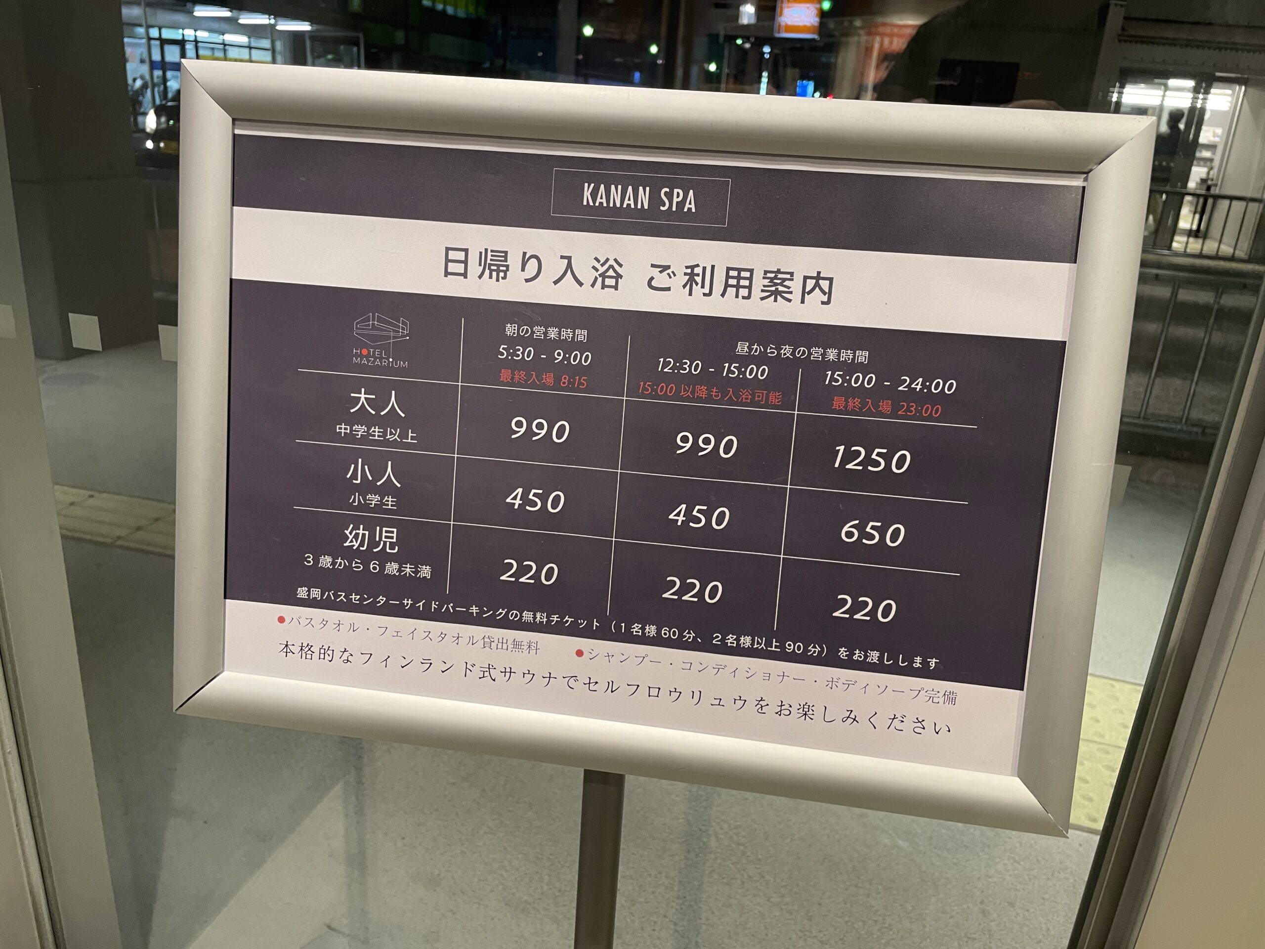 KANAN SPAの日帰り入浴案内看板｜料金表と営業時間が白地のパネルに整理され、金属スタンドに掲示された入口付近の案内表示｜岩手県盛岡市
