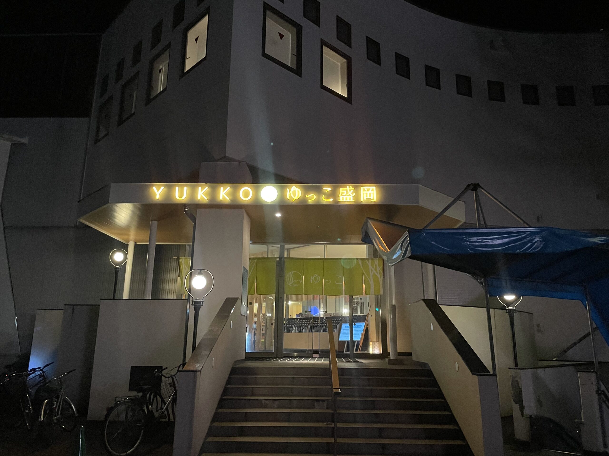 夜のSPA銭湯ゆっこ盛岡の正面入口｜黄色く照らされたYUKKOの看板と階段、ガラス張りのエントランスが見える外観｜岩手県盛岡市